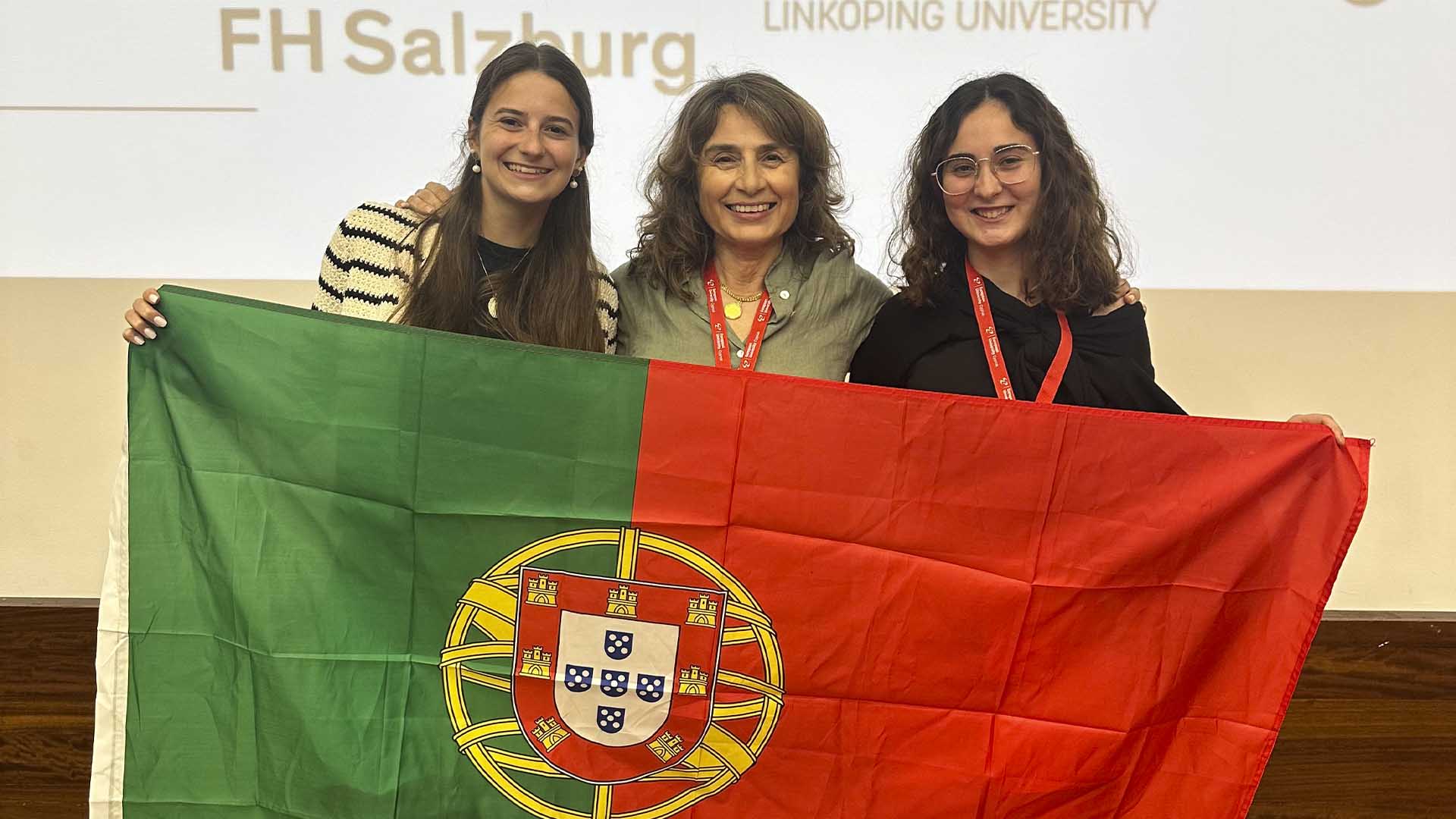 duas alunas e professora sorridentes a segurar bandeira portuguesa