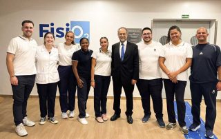 Grupo de alunos da ESSALCOITÃO com o Bastonário da Ordem dos Fisioterapeutas, posa para a fotografia durante a inauguração da Unidade de Fisioterapia Cardiorrespiratória e Sarcopenia (FisiO2). Estão num espaço interior, junto a uma parede com o logótipo “FisiO2”, e todos sorriem para a câmara.