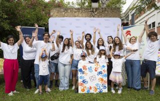 Um grupo diverso de crianças, jovens e adultos posa junto num jardim, todos a sorrir e com os braços erguidos no ar. Usam t-shirts “Dream. Speak. Live.” e seguram uma tela colorida com marcas de mãos em laranja e azul.