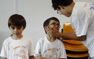 Três crianças usam t-shirts com o logótipo “Dream. Speak. Live.”. Duas delas interagem de forma carinhosa, com uma a segurar o rosto da outra e a sorrir. A terceira observa a cena com expressão calma.