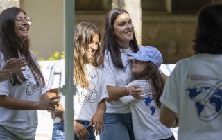 Um grupo de jovens sorri e conversa ao ar livre, usando t-shirts brancas com o logótipo “Dream. Speak. Live.”. Uma das participantes abraça uma criança com boné lilás. O ambiente é descontraído e alegre.